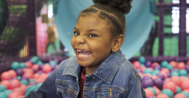 A girl in a ball pit smiling big.