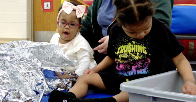 Two children sitting on the floor, one playing with metallic paper and the other playing with silver sound balls.